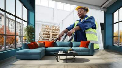 Young African American female worker of warehouse pushing forklift loader while moving along spacious workshop of factory Wall mural