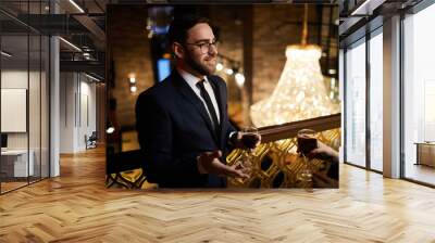 Young businessman in formalwear having red wine while talking to colleague at corporate party in classy restaurant Wall mural