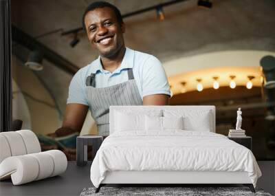 Young cheerful man in apron looking at you with toothy smile while counting money Wall mural