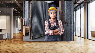 Young confident Hispanic female engineer in coveralls and protective helmet crossing arms by chest while standing in warehouse Wall mural