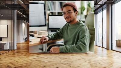 Young confident IT engineer or software developer in casualwear sitting in armchair by computer monitor and laptop and looking at camera Wall mural