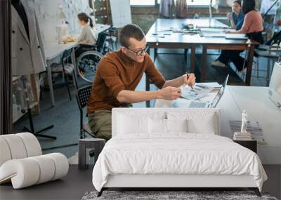 Young disable man sitting by table and working over fashion collection in large workshop Wall mural