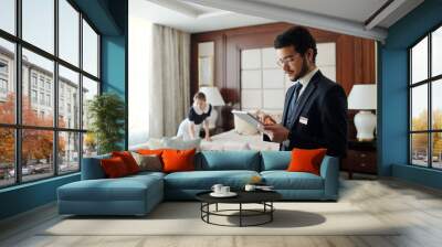 Young elegant entrepreneur in formalwear using tablet while standing in front of camera in hotel room with chambermaid on background Wall mural