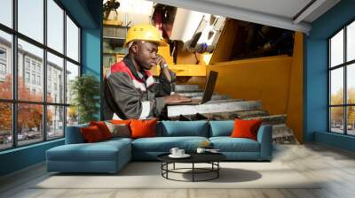 Young serious African American male technician in workwear and hardhat looking at laptop screen while looking through online repair manual Wall mural