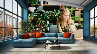 Young serious blond businesswoman with cup of coffee sitting by desk in front of laptop and networking in office with green plants Wall mural