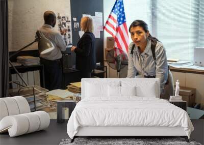 Young serious Hispanic female detective bending over workplace with documents on background of two colleagues communicating Wall mural