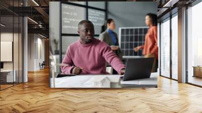 Young serious male entrepreneur or architect pressing key of laptop keyboard and looking at screen while sitting by workplace Wall mural