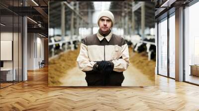 Young serious male owner of modern cowfarm in uniform standing in front of camera against two cowsheds with livestock and looking at you Wall mural