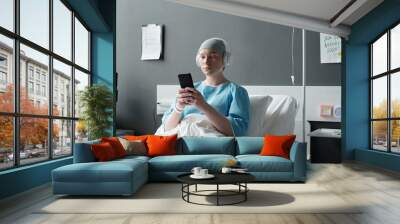 Young sick woman with oncological disease using smartphone while sitting in bed in hospital ward after chemotherapy treatment and having rest Wall mural