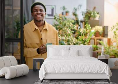 Young smiling African American businessman with cup of tea or coffee looking at camera while standing against his workplace in office Wall mural