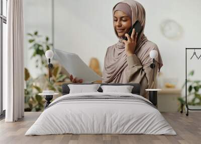 Young smiling businesswoman in hijab speaking on mobile phone and reading contract terms and conditions to client in home environment Wall mural