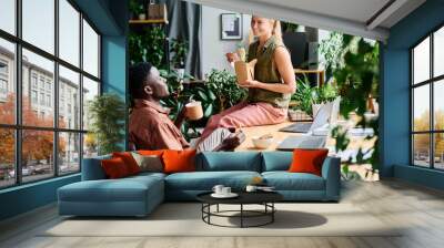 Young smiling female office manager eating wok and talking to African American male colleague sitting by workplace among green plants Wall mural