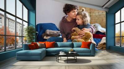 Young woman giving hug to her cute little son with brown soft teddybear while both sitting on sleeping place prepared for refugees Wall mural