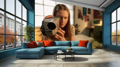 Young woman holding retro handheld video camera in cozy indoor setting with bed and posters in background. Person engaging in creative activity involving digital media Wall mural