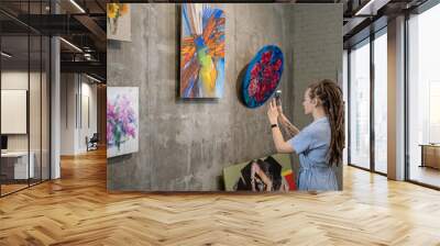 Young woman standing in front of the wall with modern art and using her mobile phone to make photos Wall mural