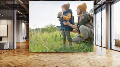 Young woman with backpack squatting by her little son with binoculars on trip Wall mural