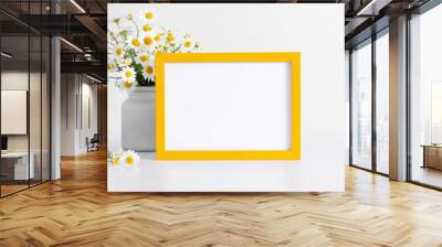 White chamomile flowers in vase and yellow photo frame on white table. Wall mural