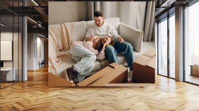 Happy young family couple man and woman relaxing on sofa after moving cardboard boxes to new estate home apartment. Wall mural