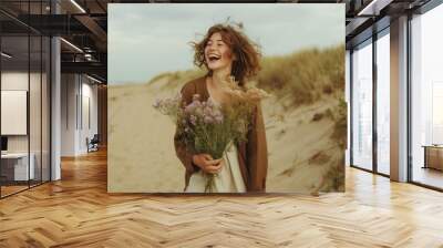 young female laughing, holding wildflowers, walking on a sandy beach Wall mural