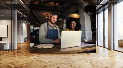 Male and mixed race female coffee shop owners smiling at laptop in coffee shop. Wall mural