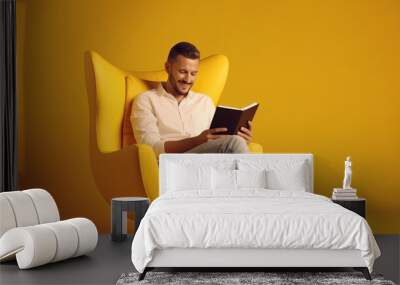 Full-length image of a perceptive, happy man wearing jeans and an armchair, reading news on a tablet against a yellow background Wall mural