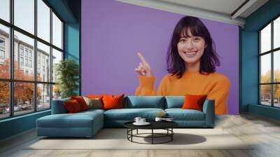 Smiling Japanese woman wearing a bright orange sweater. She is pointing with her right hand, standing against a solid purple background. Wall mural