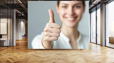 Young woman smiles with a thumbs-up gesture in a neutral background during a cheerful moment of positivity and encouragement. Wall mural