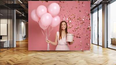 Beautiful smiling woman in dress and birthday hat holding gift box and pastel pink air balloons and falling confetti against pink background. cute happy young girl celebrating birthday party. Wall mural