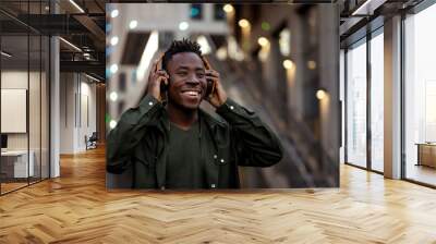 smiling african-american man in stylish jacket in wireless headphones listening music on the street of the evening city. space for text Wall mural