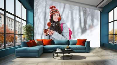 Tourist brunette girl wearing grey jacket, hat and mittens standing with thermo cup and considering the winter landscapes in the winter cold forest Wall mural