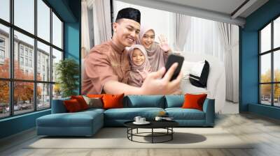 Young Indonesian family doing video call to other family and relatives with greetings gesture to the phone in happy during Eid mubarak in the living room at home. Wall mural