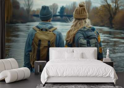 A man and a woman in knitted hats stand with their backs to the viewer. They wear flannel shirts and plaid backpacks. They look at the river and bridge in the background. Wall mural