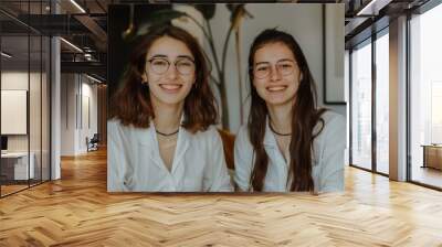 Two young women wearing glasses sit next to each other and smile at the camera. Best friends. Wall mural