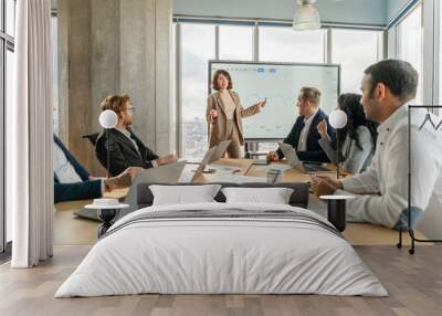 A businesswoman stands in front of a large screen and points to a graph as she presents data to a team of colleagues seated around a table in a modern office. Wall mural