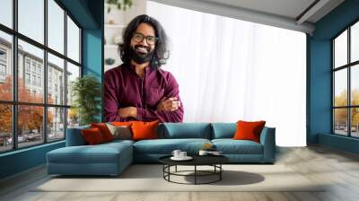 A cheerful Indian man with long curly hair and glasses stands with arms crossed, exuding confidence and warmth. The bright room is decorated with natural light, copy space Wall mural