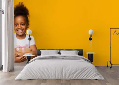 A cheerful young black girl stands confidently, her curly hair styled high as she smiles brightly. Her hands are crossed in front of her while she enjoys this lively moment against yellow backdrop. Wall mural