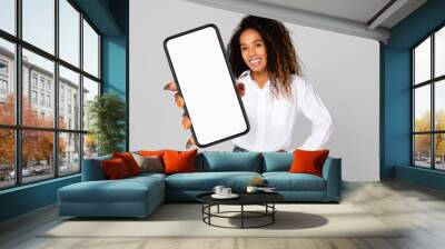 A cheerful young black woman with curly hair is holding a smartphone with a blank screen towards the camera. Wall mural