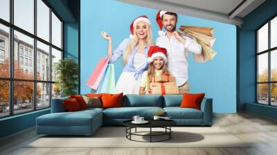 A family of three, wearing Santa hats, smiles for the camera while holding Christmas presents and shopping bags. The parents are standing behind their daughter, who is holding a stack of wrapped gifts Wall mural