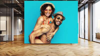 A happy black couple is pictured against a bright blue background. The woman is wearing a red swimsuit and a straw hat, while the man is shirtless and wearing a straw hat and sunglasses. Wall mural