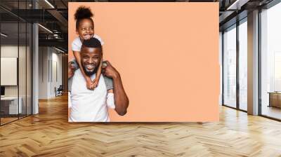 A happy black father and daughter pose for a picture against a peach-colored background. The little girl is sitting on her fathers shoulders, and both are smiling brightly at the camera, copy space Wall mural