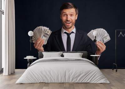 A man in a suit holds a stack of cash in each hand in front of a dark background. Wall mural