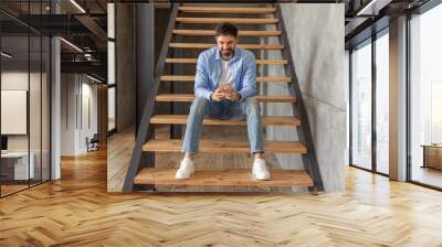 A man sits on a set of wooden stairs in a modern home. He wears a blue shirt, white t-shirt, blue jeans, and white sneakers. He is looking down at his phone and smiling. Wall mural