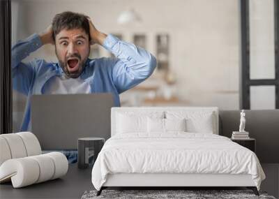 A man sitting on a couch at home is holding his head in frustration while staring at his laptop computer. He shows a mix of shock and stress during a challenging work moment. Wall mural