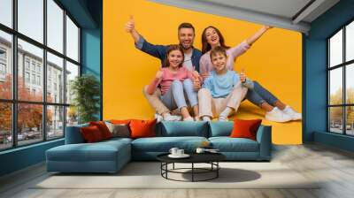 A smiling family consisting of a mother, father, daughter, and son are sitting close to each other against a vibrant yellow backdrop, all giving a thumbs-up sign in a show of happiness and unity. Wall mural