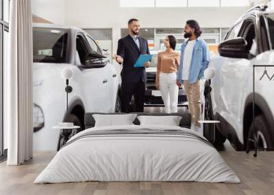 A well-dressed salesman is presenting information to Indian couple in a car dealership showroom. The couple appears interested as they stand between two new vehicles Wall mural