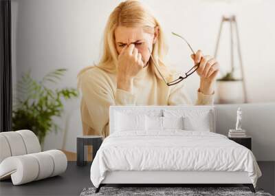 A woman sits at a desk, rubbing her eyes and holding her glasses. She looks tired and may be experiencing eye strain from working on her laptop. Wall mural