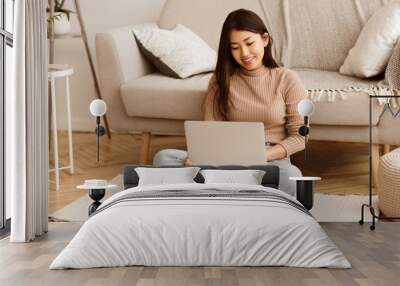 A woman sits on a rug in a living room, using a laptop computer. She is smiling and appears to be focused on her work. Wall mural