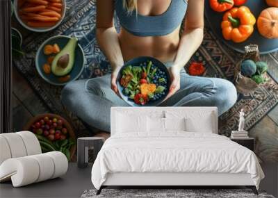 A woman sits on a rug with her legs crossed while holding a bowl of fresh salad. She is eating a healthy meal with vegetables and fruits. Wall mural