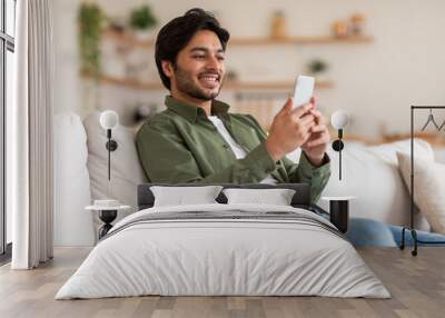 A young Arab man with a beard sits on a white couch while holding and looking at a white smartphone. He is wearing a green shirt and blue jeans. Wall mural