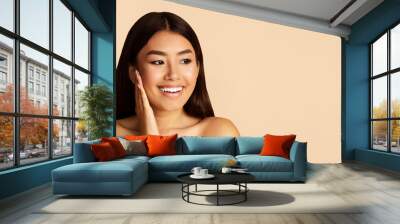 A young Asian woman with long black hair smiles widely as she touches her cheek with her hand. She is looking off to the side, and the background is a soft peach color, copy space, panorama Wall mural